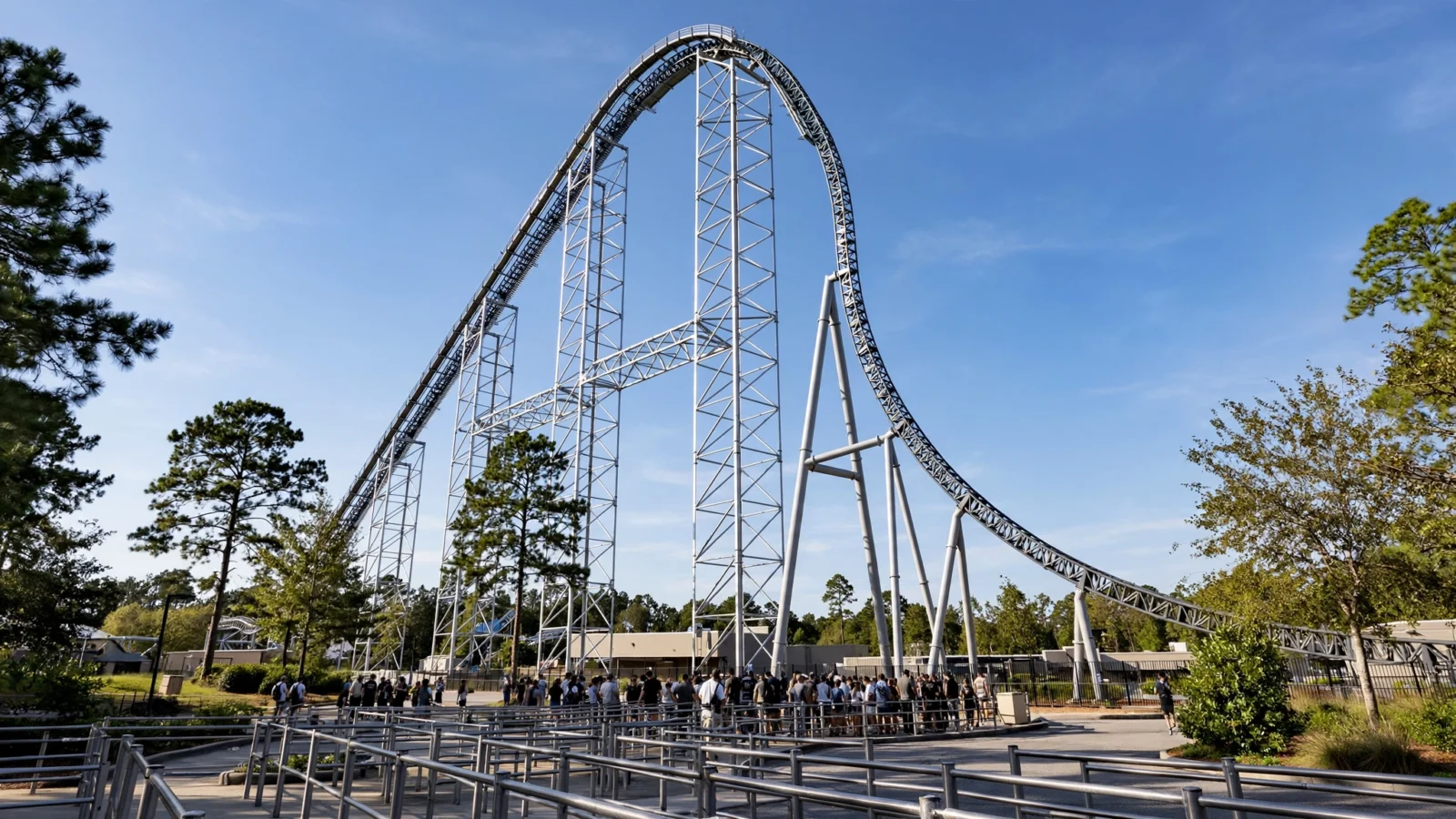 A tall steel roller coaster with a steep first drop, viewed from the ground, with visitors waiting in a queue area below.