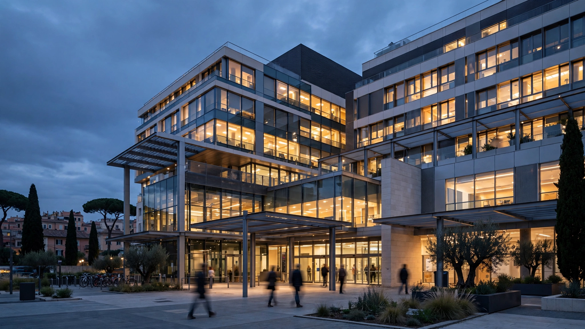 A modern hospital building in Rome lit up at dusk, with blurred figures walking near the entrance