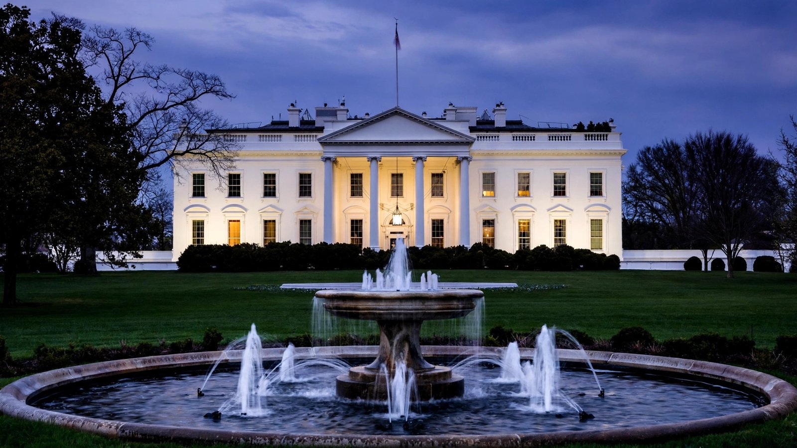 The White House facade with warm lights glowing in the windows at dusk, seen across the lawn