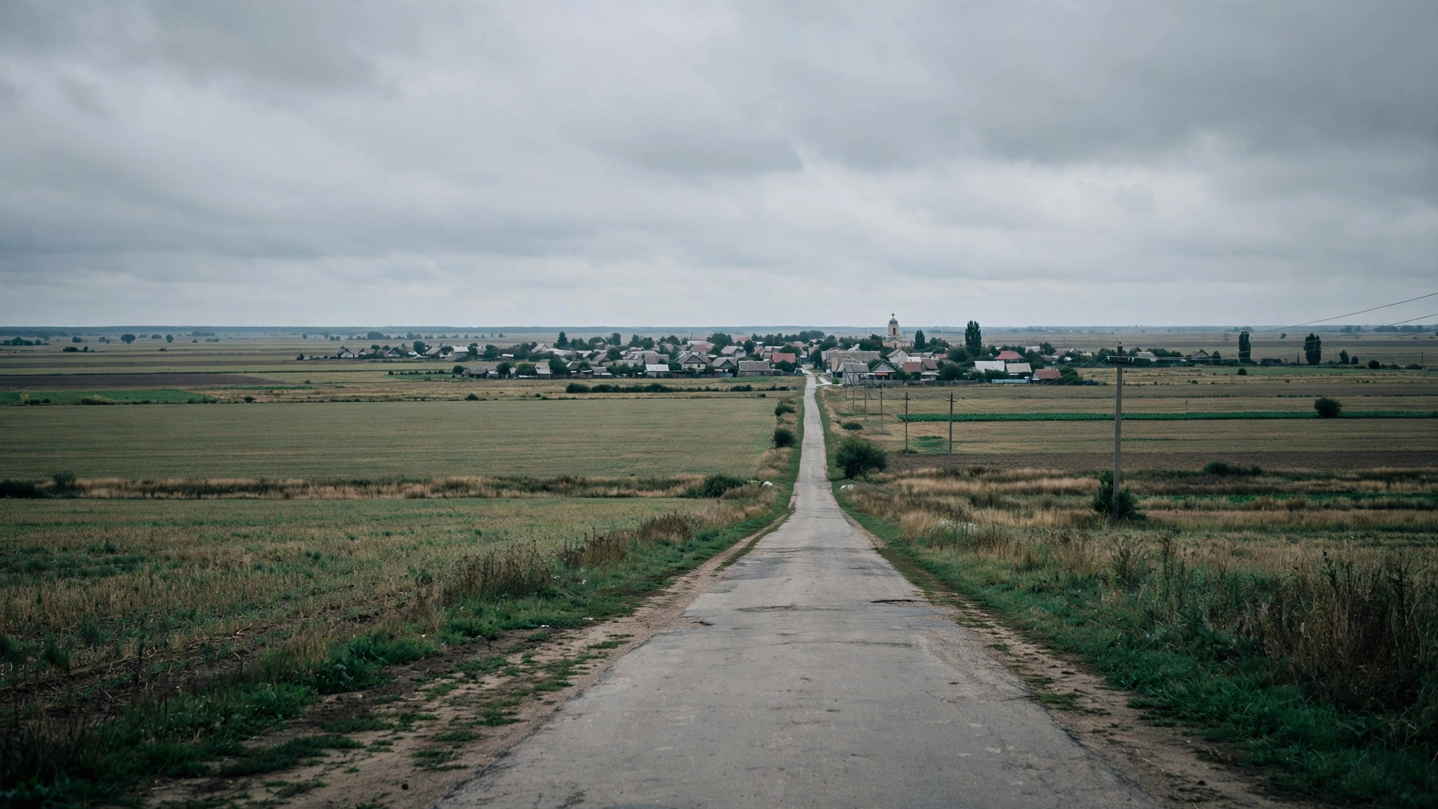 A straight rural road leading through flat farmland toward a small town under an overcast sky