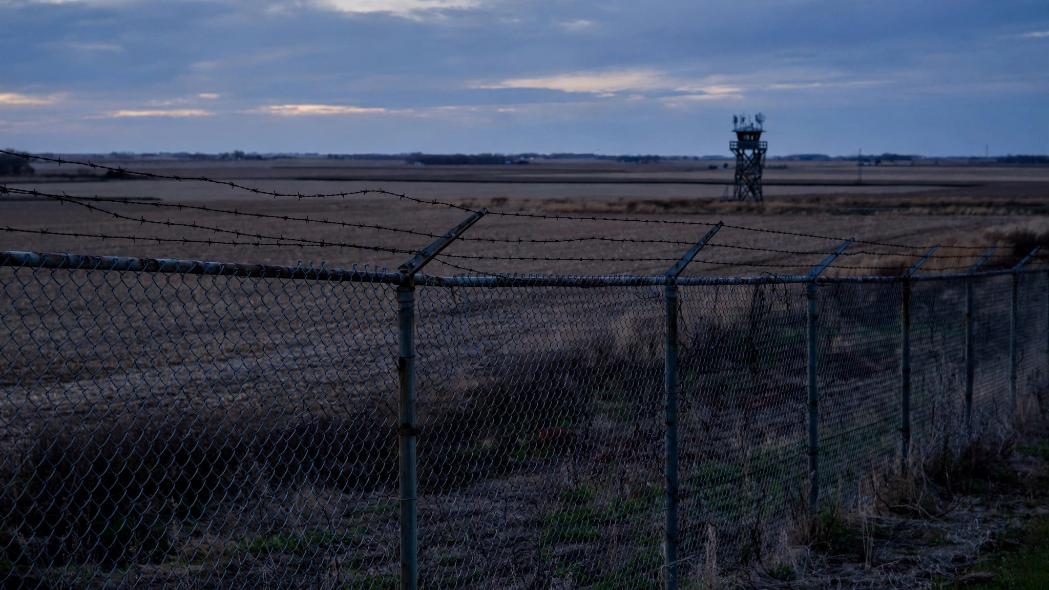 A chain-link fence topped with barbed wire at dusk with a guard tower and flat farmland in the distance