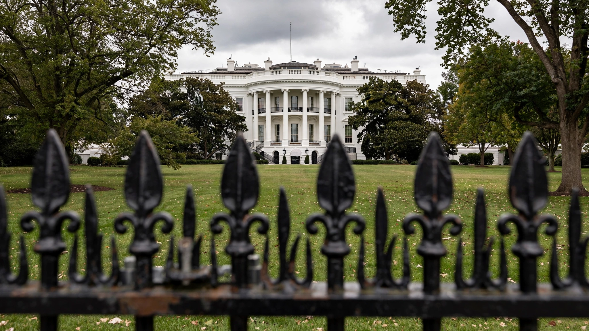 The White House seen across the lawn through a black iron fence, with trees on either side.