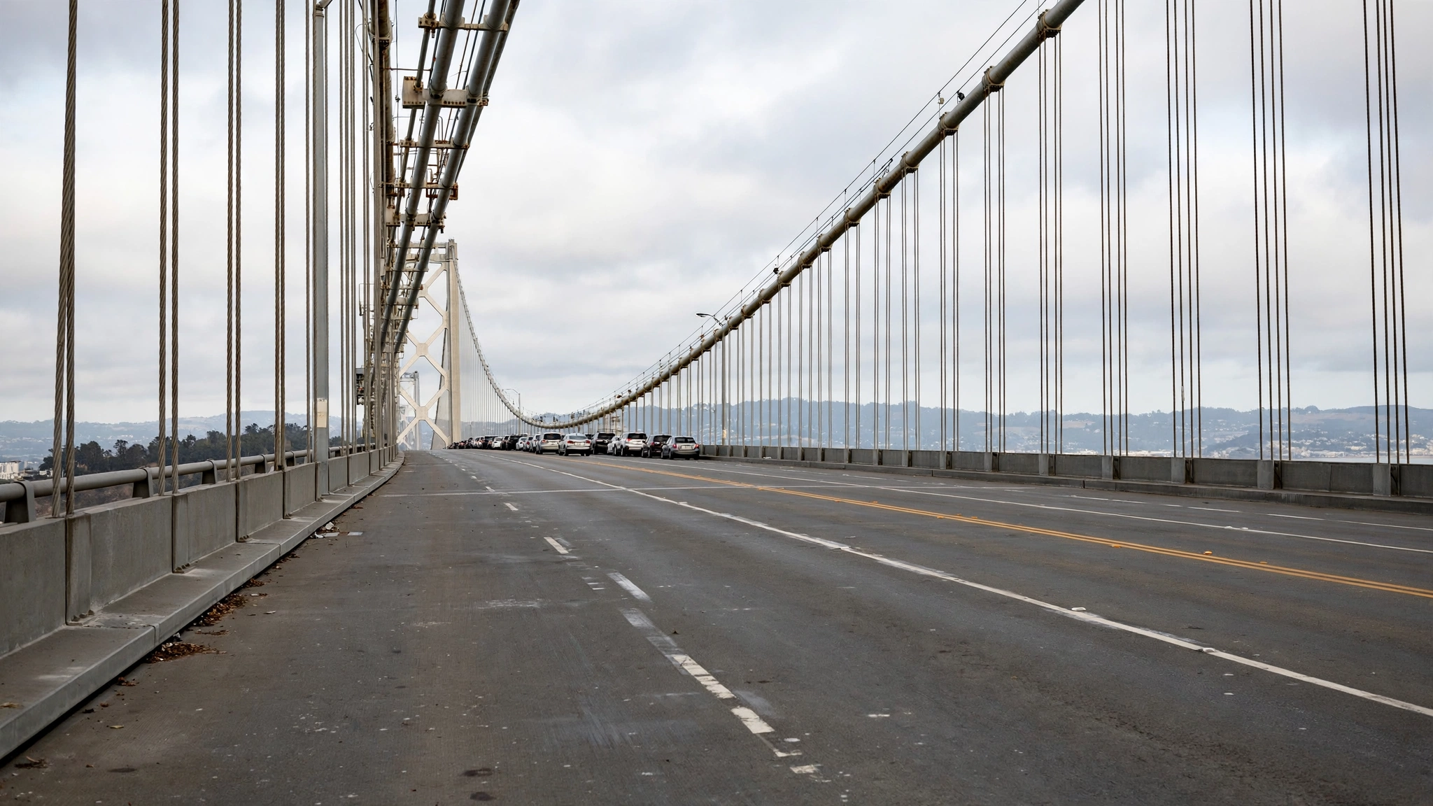 An empty bridge roadway with suspension cables overhead and a line of cars in the distance