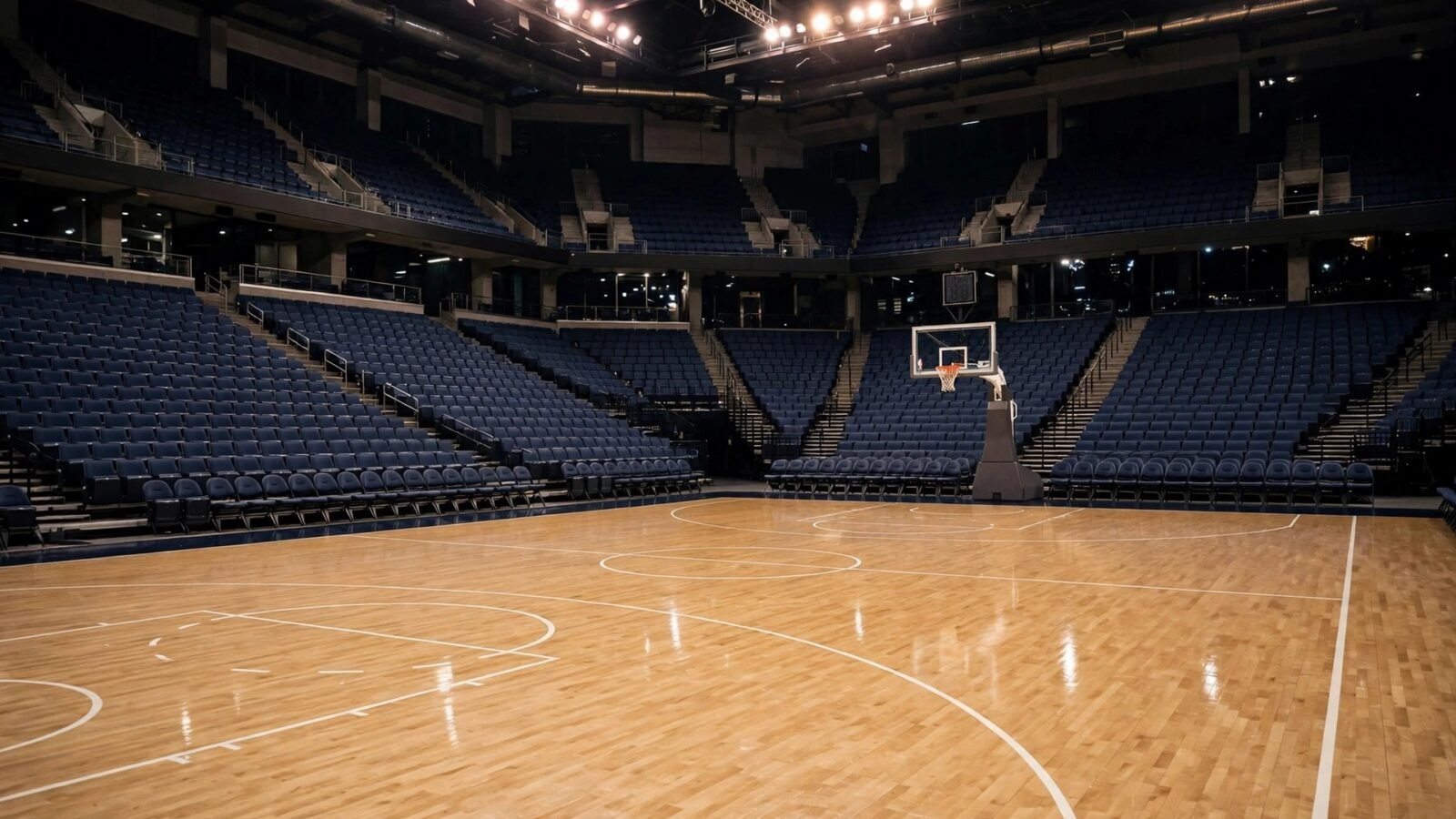 An empty basketball arena at night with a polished wooden court, a hoop and rows of blue seats under bright lights