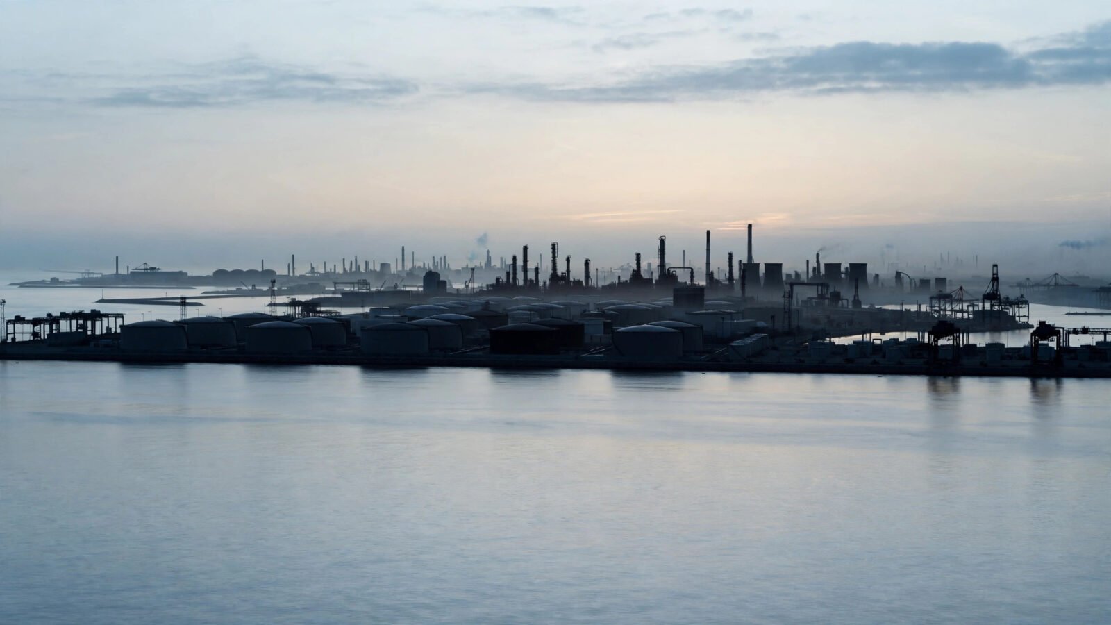 Oil storage tanks and refinery towers along a coastline at dawn, seen across calm water.