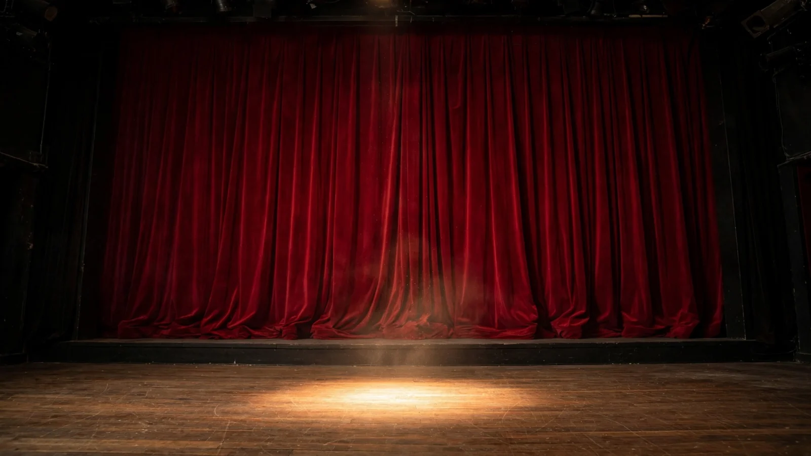 A closed red velvet theater curtain with a single spotlight on a wooden stage
