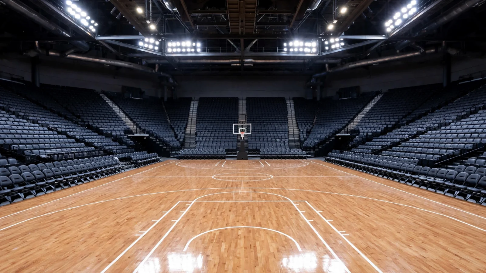 An empty indoor basketball arena with a polished hardwood court, a hoop, and bright lights above rows of seats.