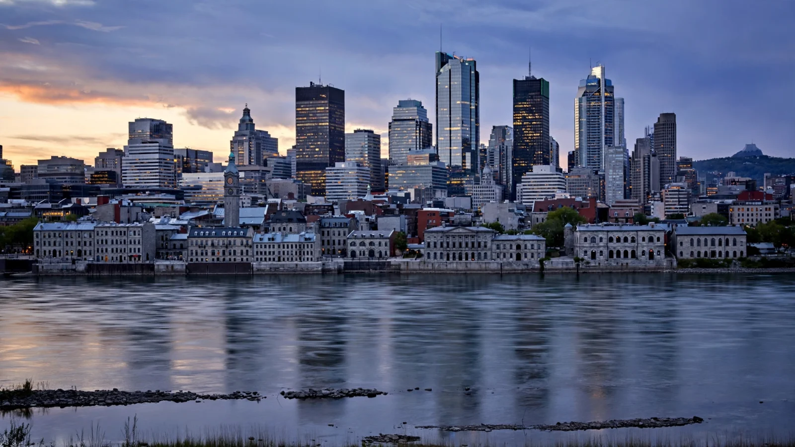 Montreal skyline at dusk seen across a river, with office towers behind historic waterfront buildings