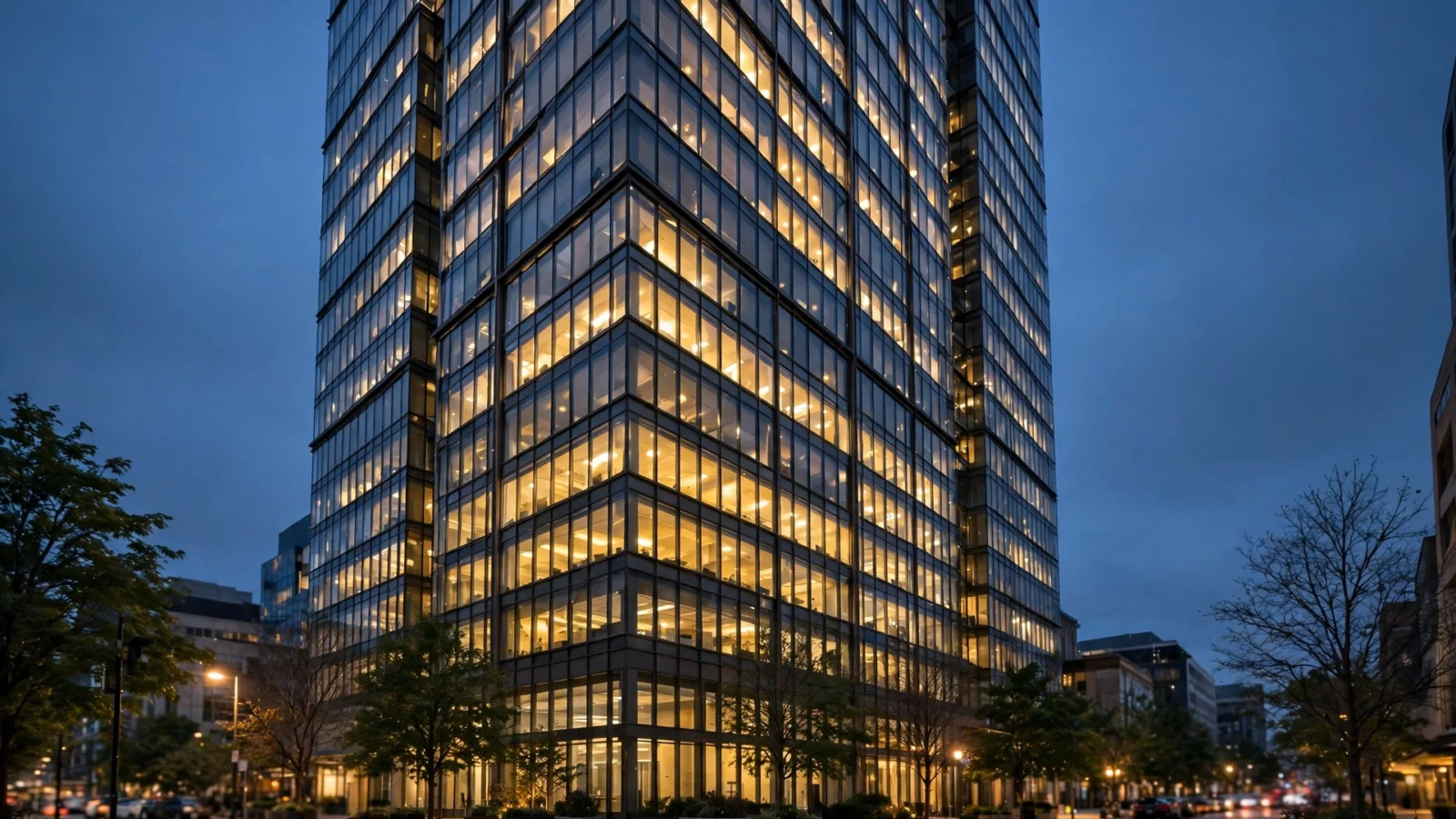 A glass office tower with lit windows at dusk, seen from a street corner