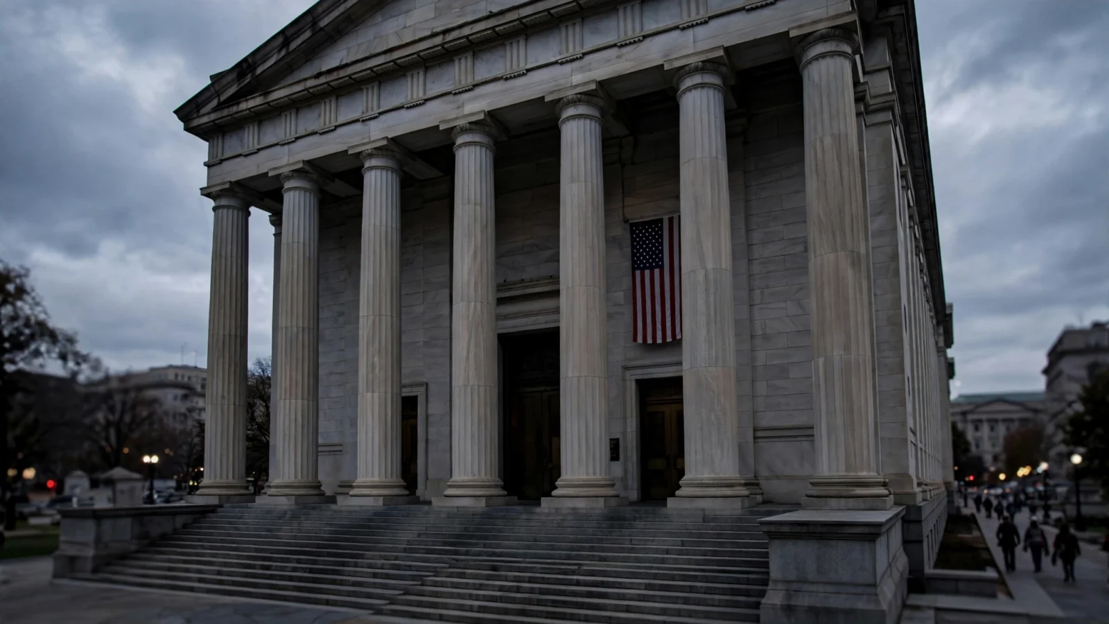 The tall stone columns and wide steps of a neoclassical federal courthouse in the United States under gray clouds, with a United States flag hanging beside the entrance and a few distant pedestrians out of focus