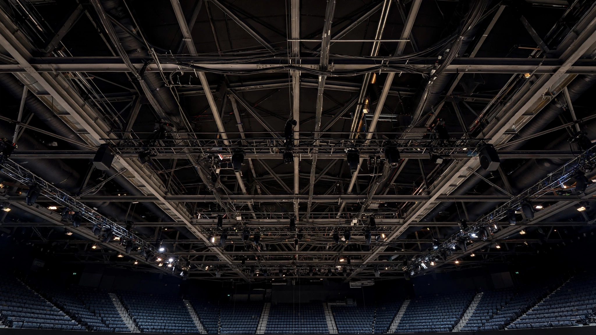 Looking up at steel rafters, cables and lighting rigs above empty seats inside an indoor sports arena