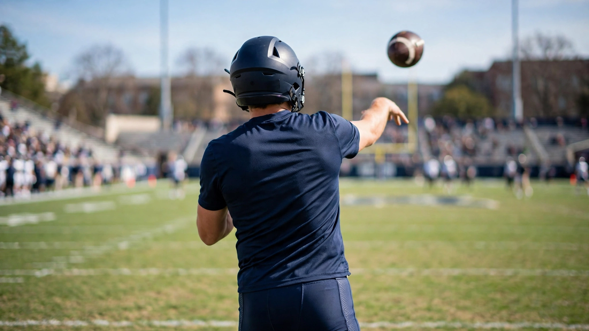 A football player seen from behind throwing a pass on a grass field with stands in the background