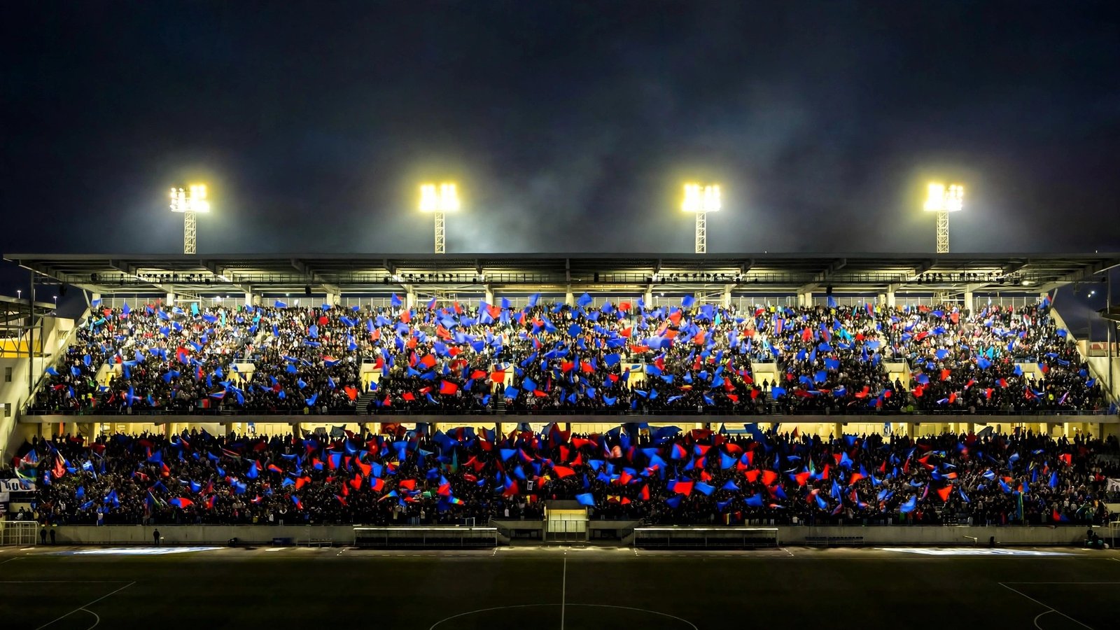 A stadium stand filled with spectators waving blue and red flags under floodlights