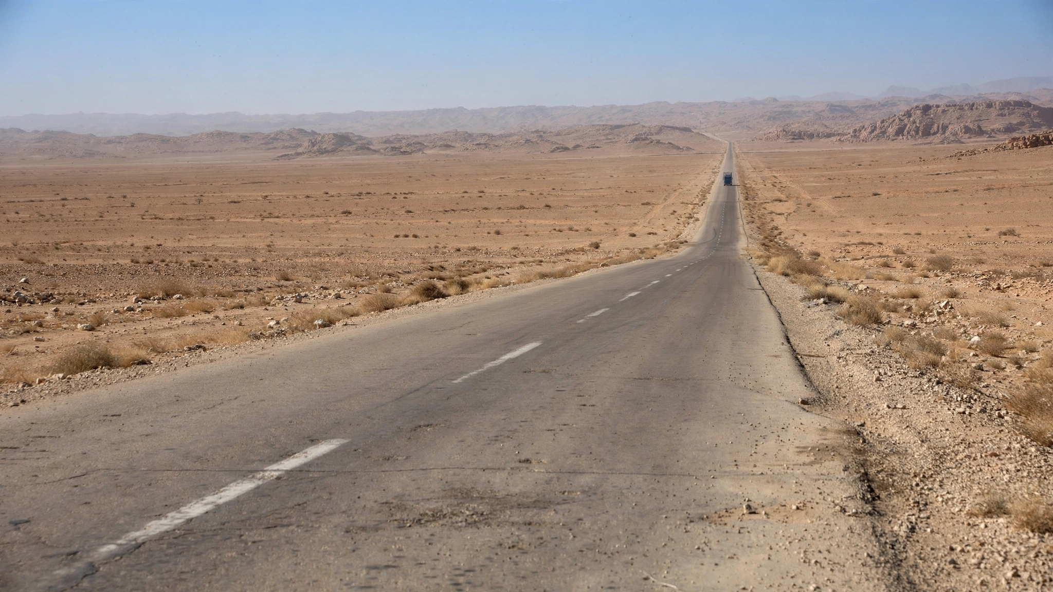 A paved road running straight through flat desert land toward distant hills, with one truck far ahead