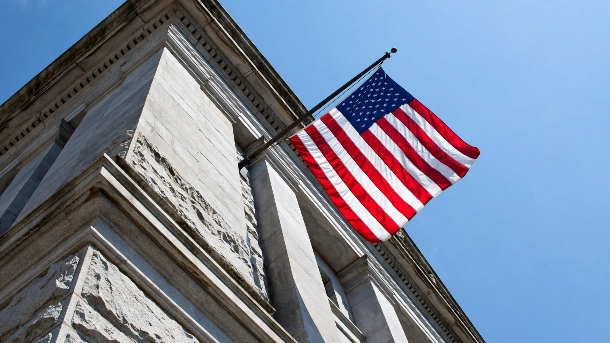 An American flag hanging from the stone corner of a government building against a clear sky