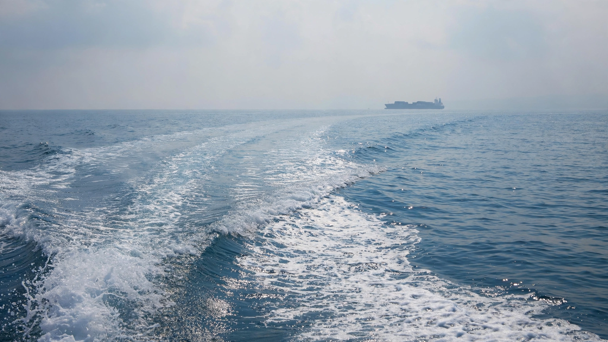 A boat wake trailing across blue sea water with a distant cargo ship on the horizon
