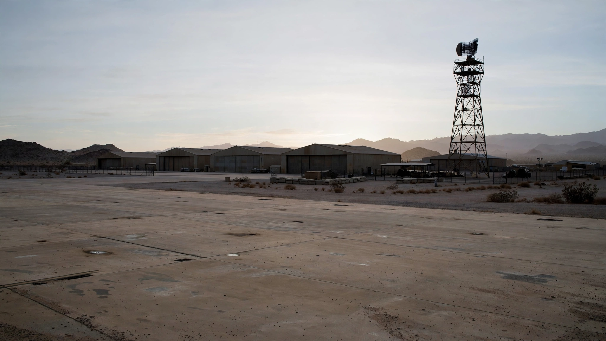 A desert air base at dawn with low hangars, an empty concrete tarmac, and a tall radar tower.