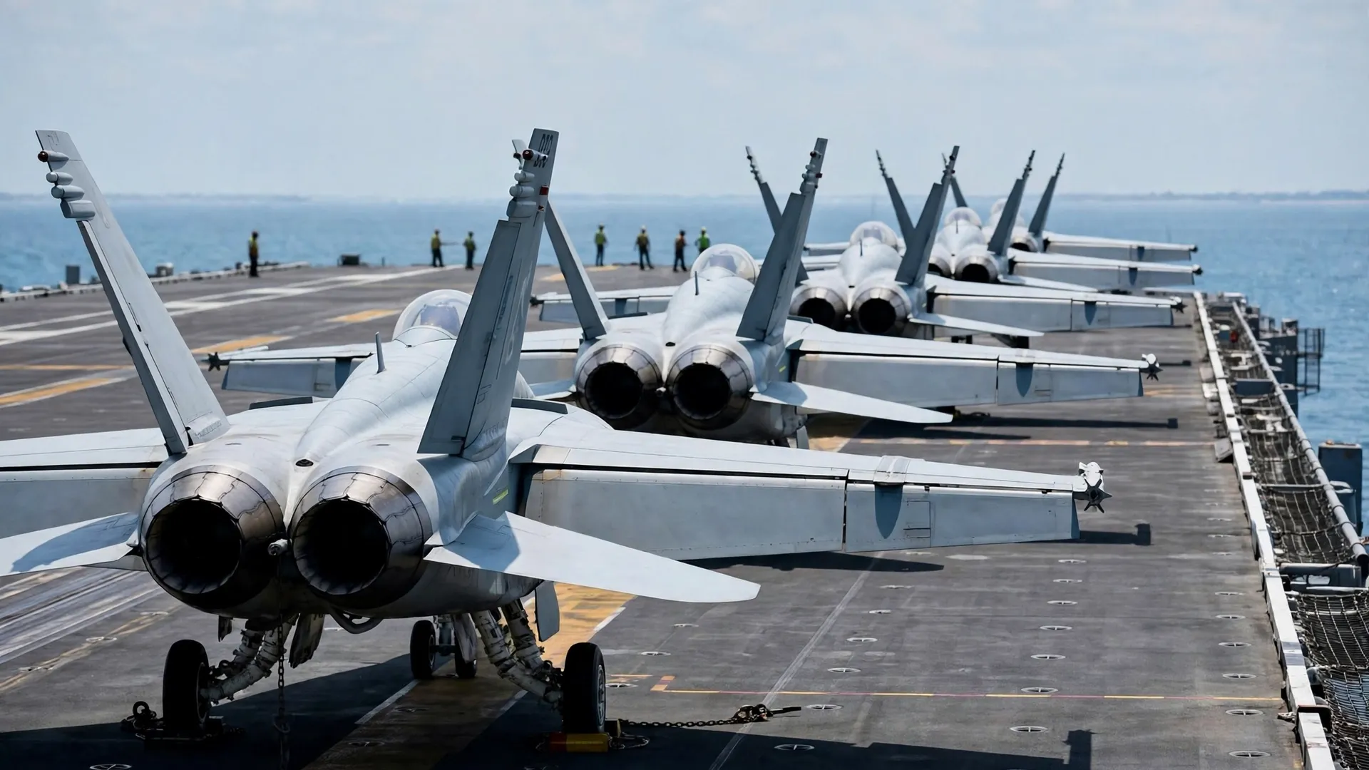 Fighter jets parked in a row on an aircraft carrier flight deck, seen from behind, with a few crew members standing in the distance.