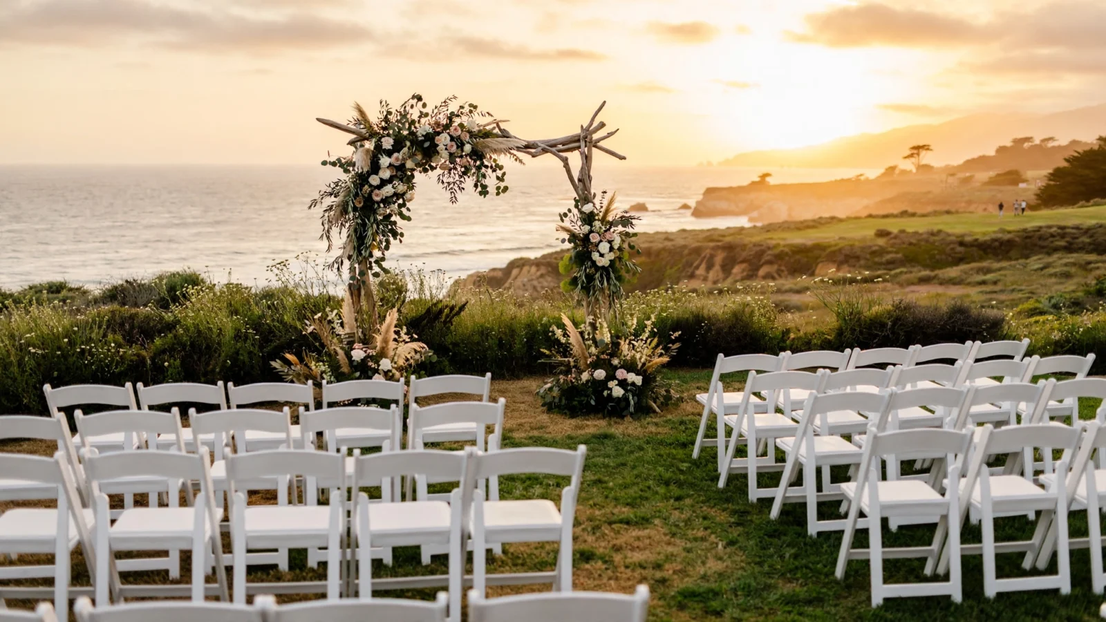 An outdoor wedding ceremony setup with white chairs and a floral arch on a California coastal bluff at sunset.