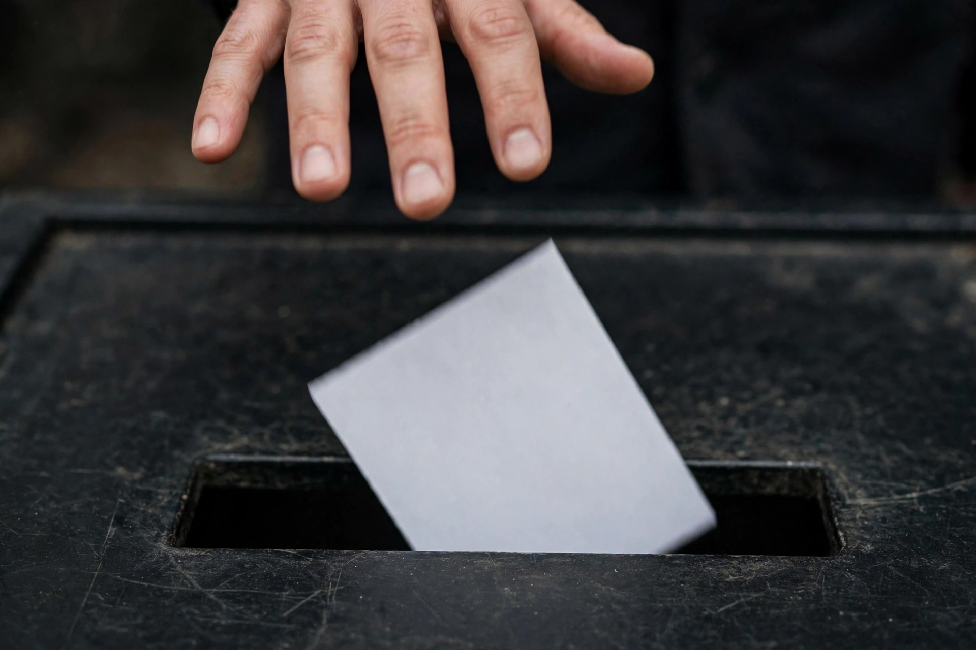 A hand placing a blank white paper ballot into the slot of a dark ballot box.