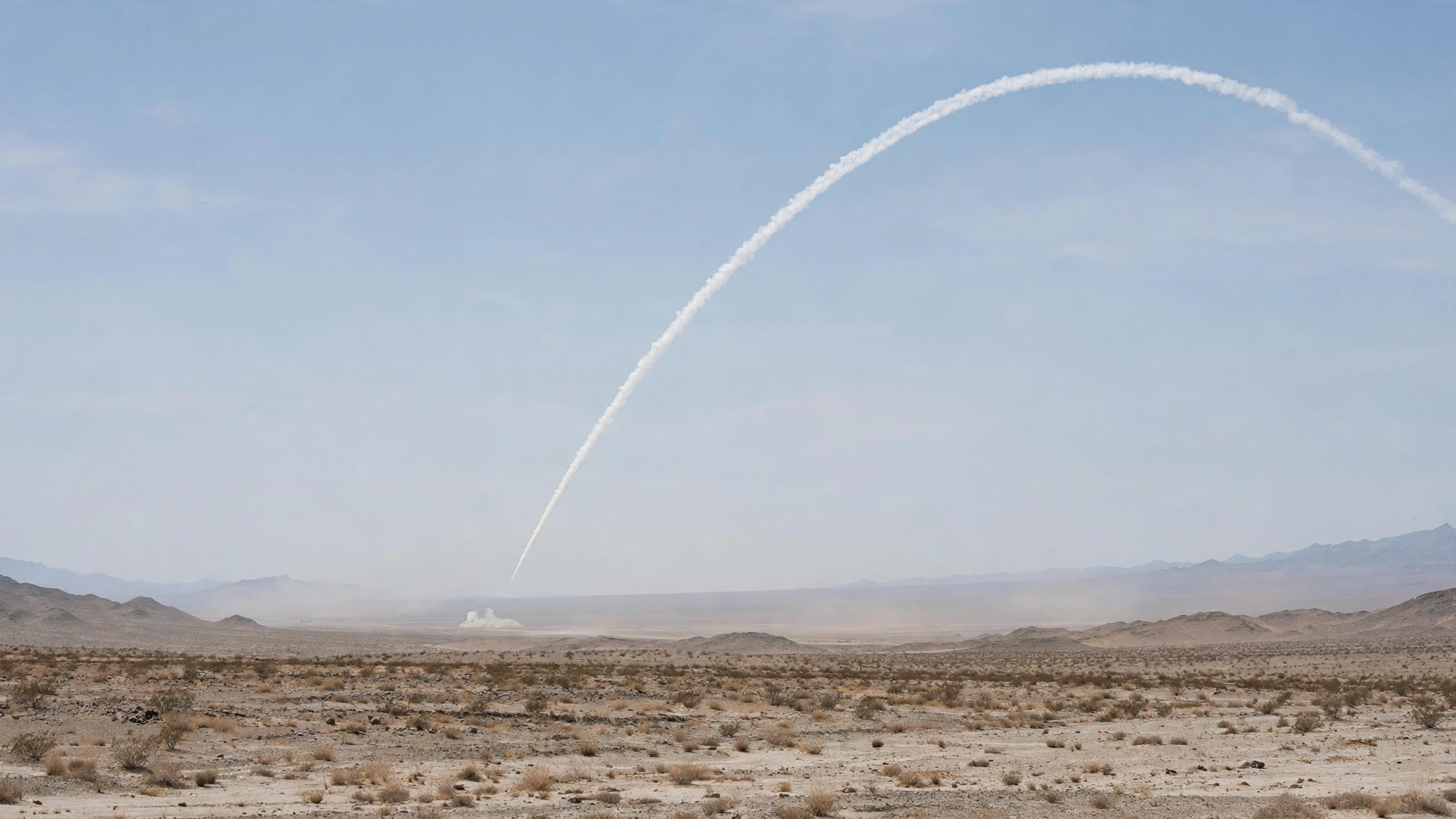 A white contrail from a distant missile test arcing across the sky over a desert