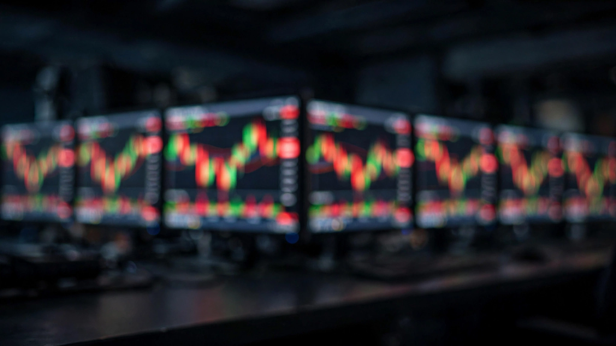Blurred monitors in a dark trading room glow with red and green stock charts.