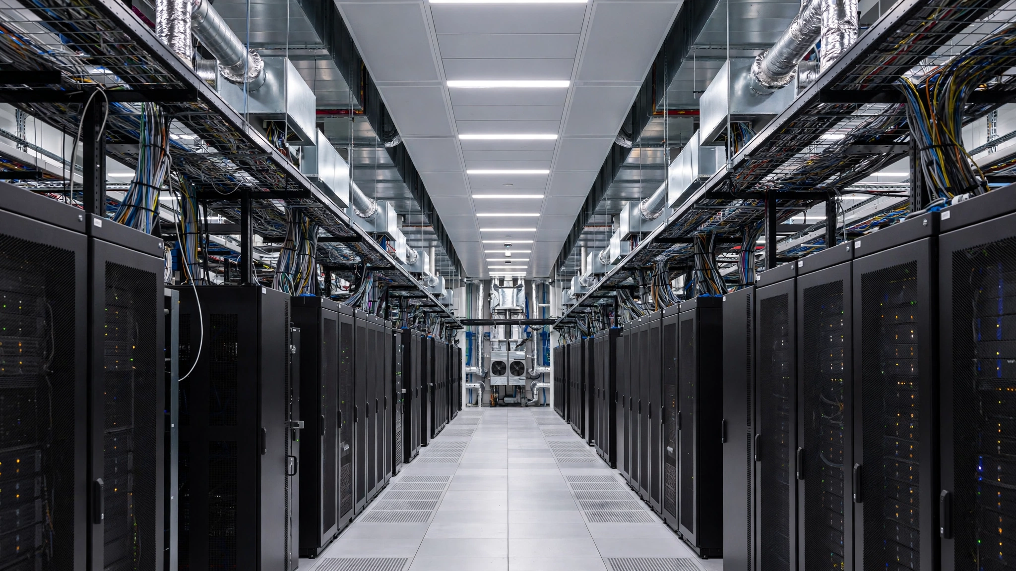 A long aisle between rows of black server cabinets with cables and cooling pipes overhead in a data hall