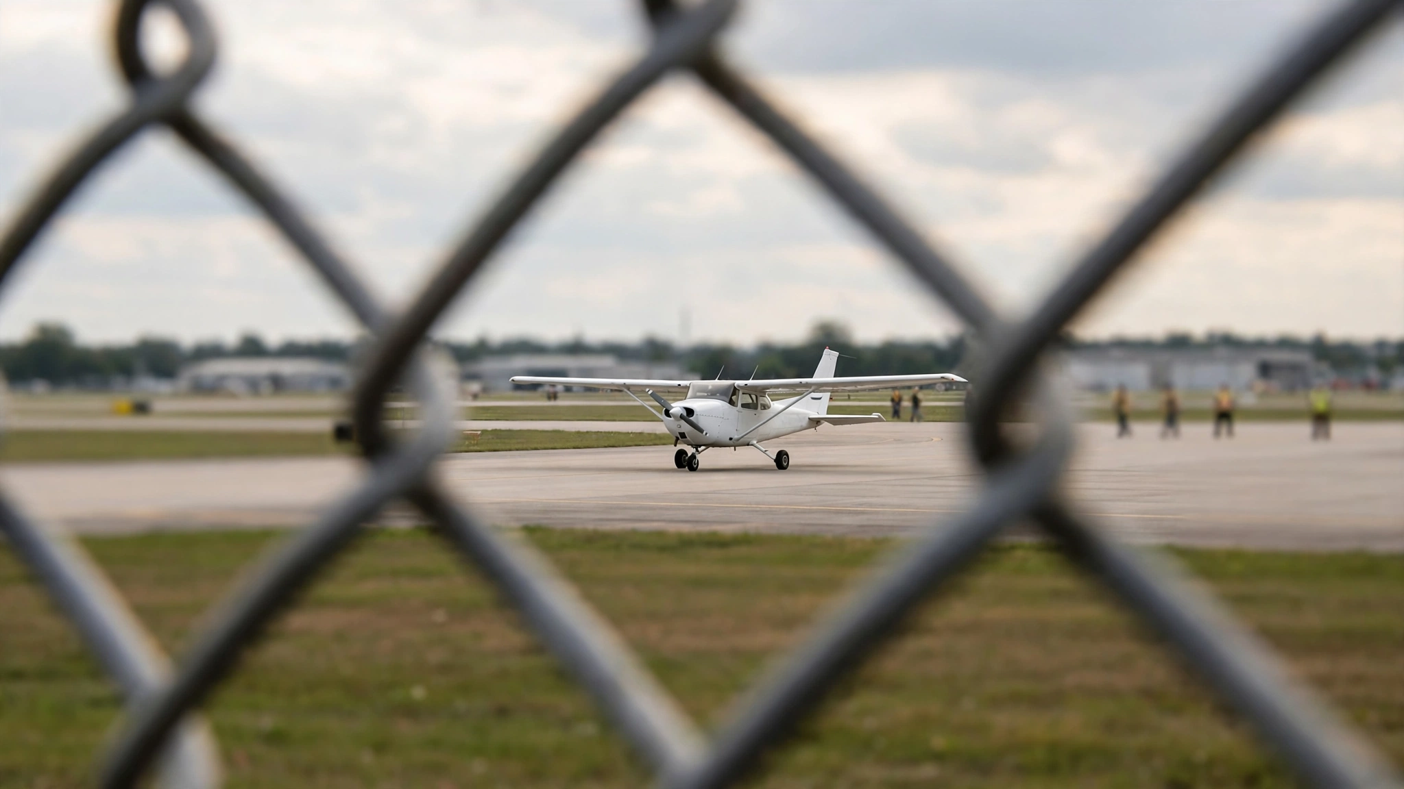 A small white propeller aircraft on a runway seen through a chain-link fence