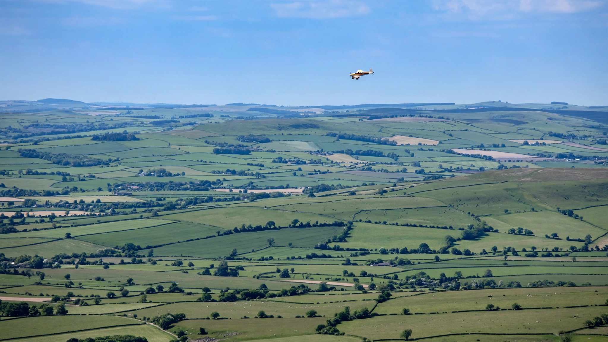 A small propeller aircraft flying high over green fields and rolling hills in daylight