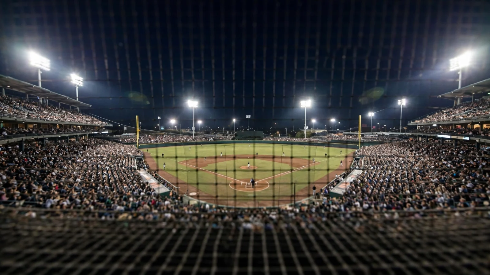Night baseball stadium filled with fans under bright floodlights, viewed through protective netting behind home plate