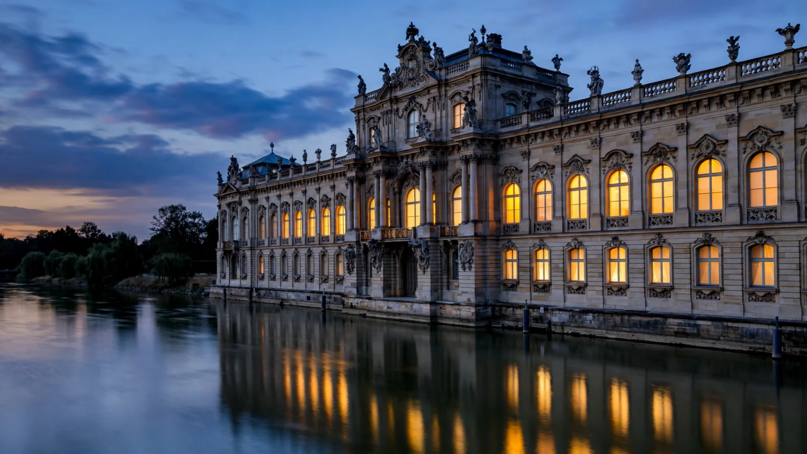 A baroque imperial palace with lit windows beside a river at dusk, reflecting in the water