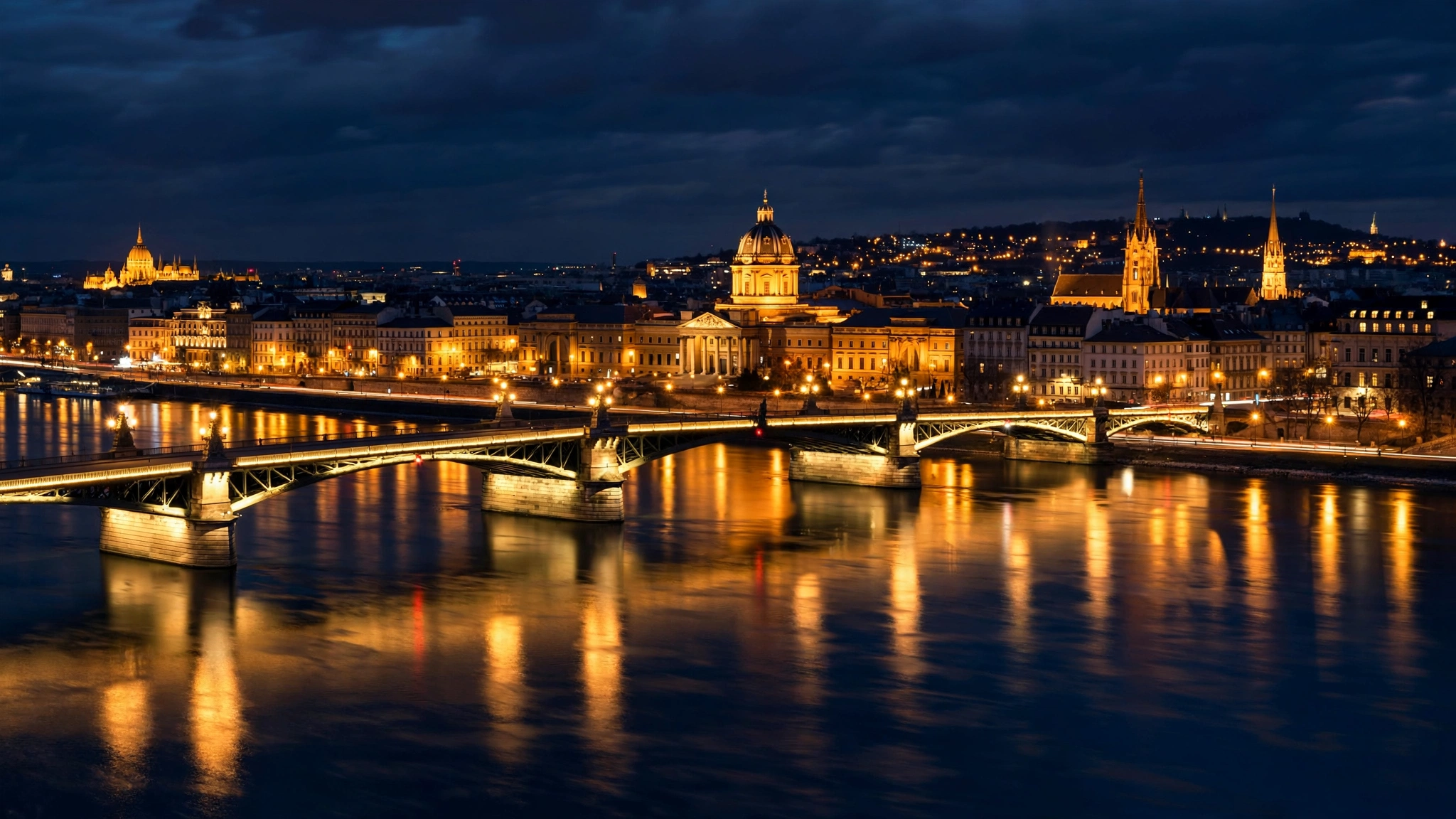 A European capital city skyline at night with an illuminated bridge crossing a river