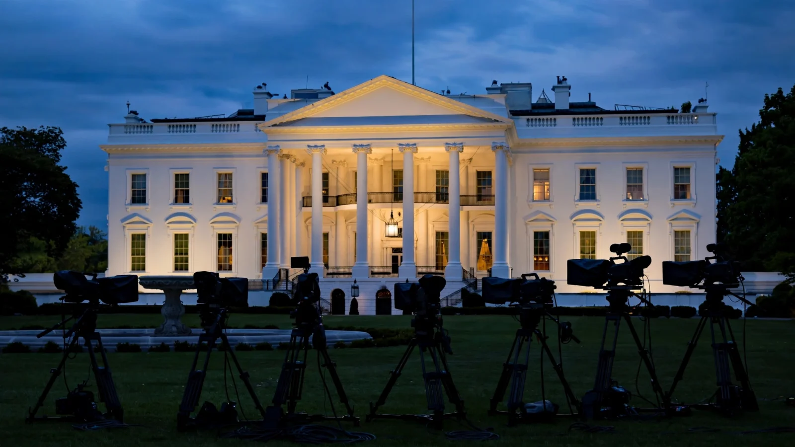 The White House north facade illuminated at dusk, with television cameras on tripods set up on the lawn in the foreground