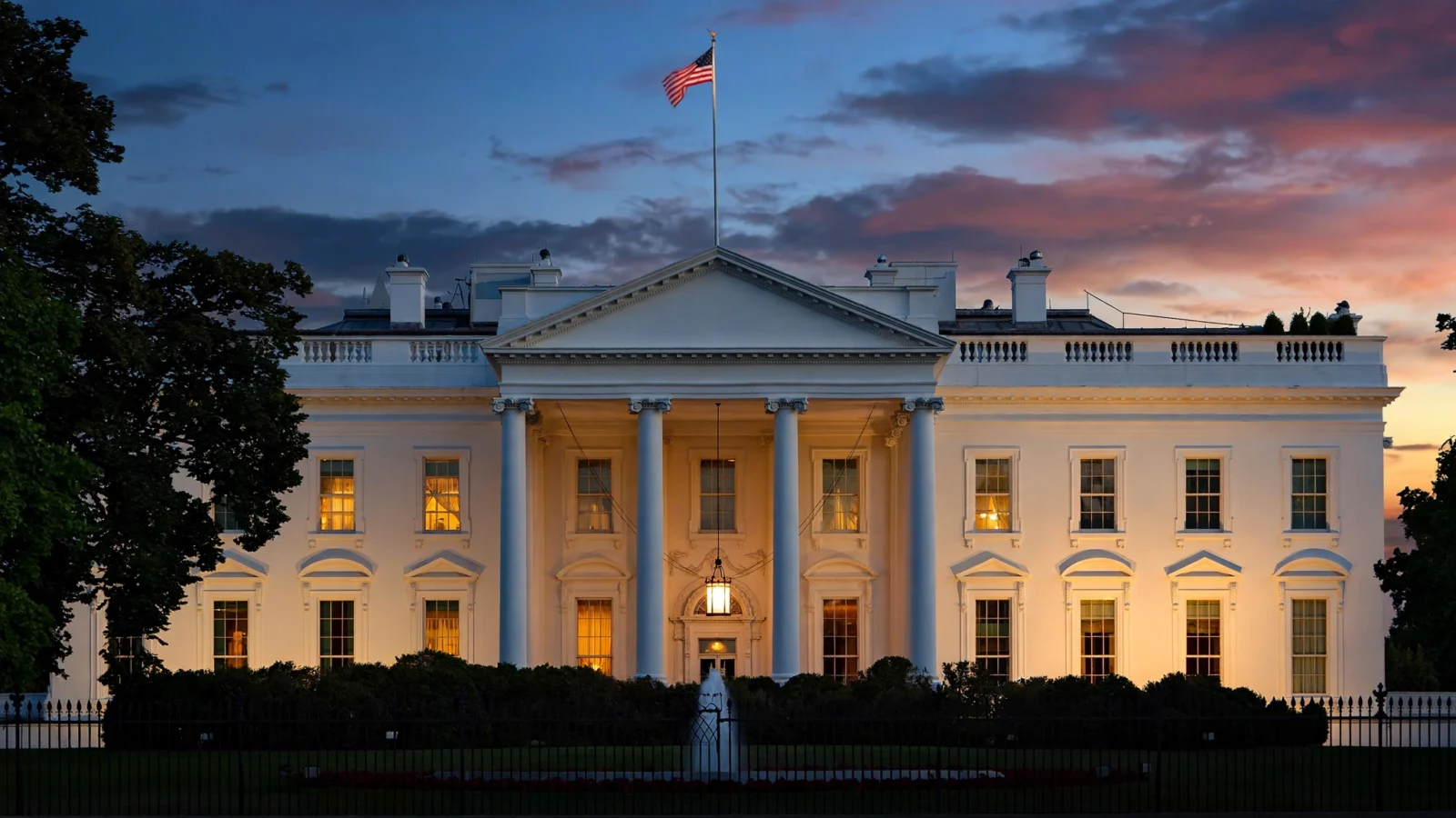 The White House exterior at dusk with the American flag flying above the lit facade