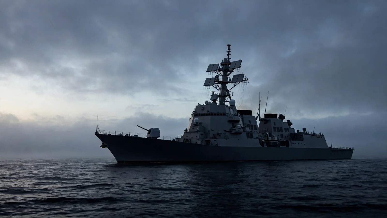 A naval warship at sea under overcast skies, seen in profile against a dark dawn horizon