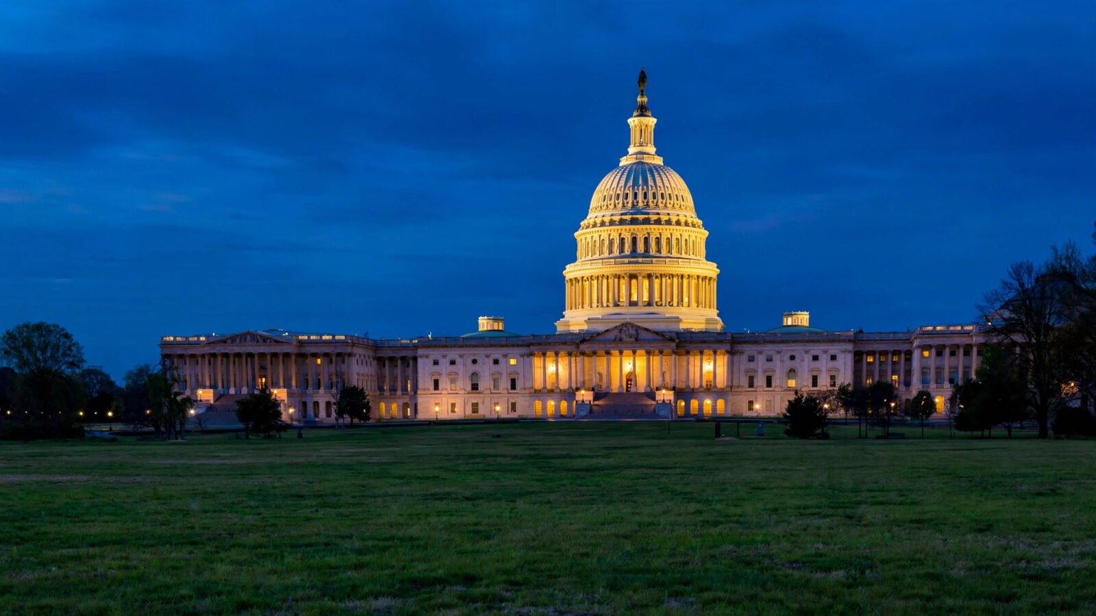 The U.S. Capitol building at dusk, its dome lit against a deep blue sky, viewed across an empty lawn