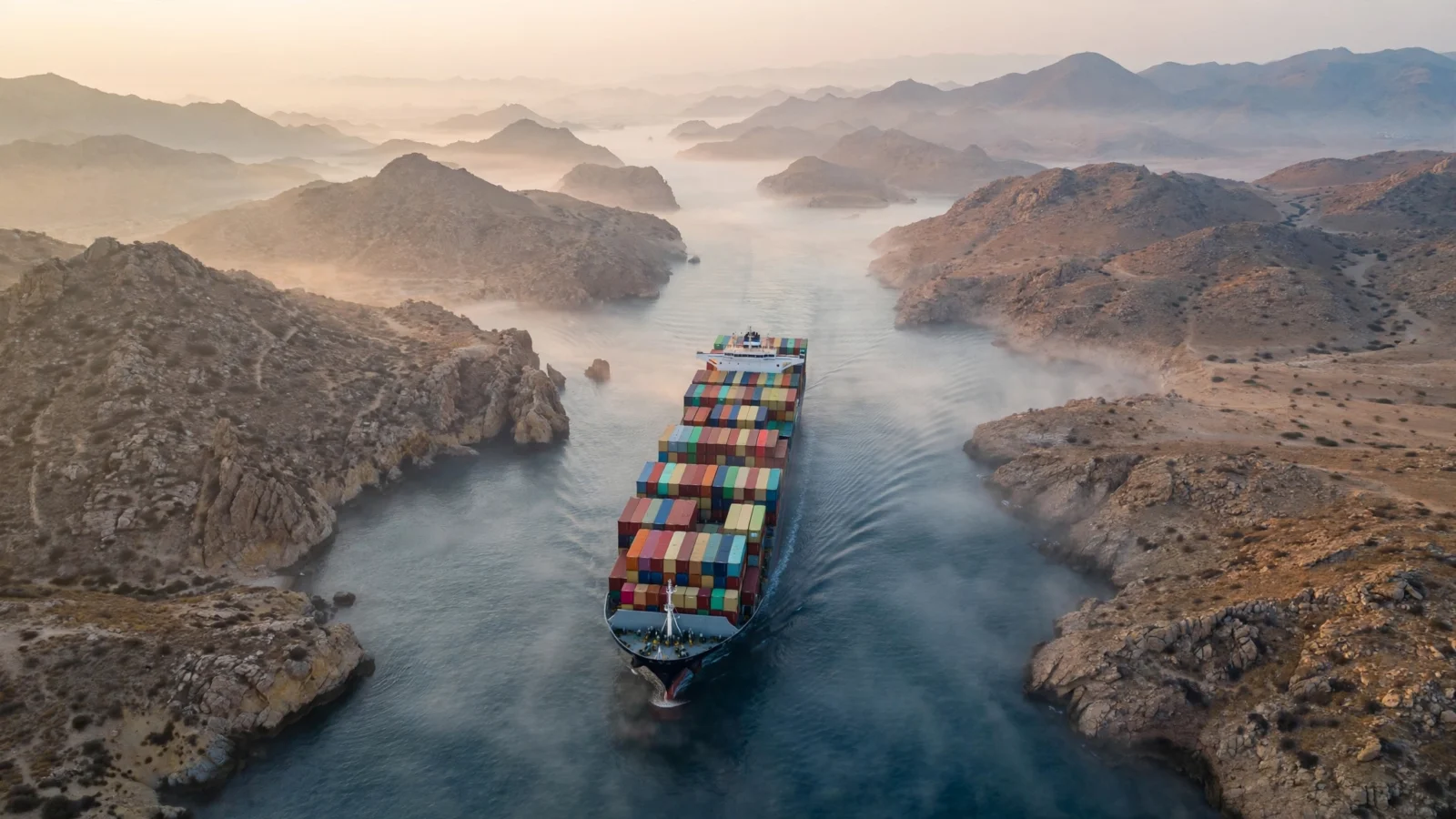 A container ship with stacked cargo containers sailing through a narrow strait between rocky coastlines at dawn