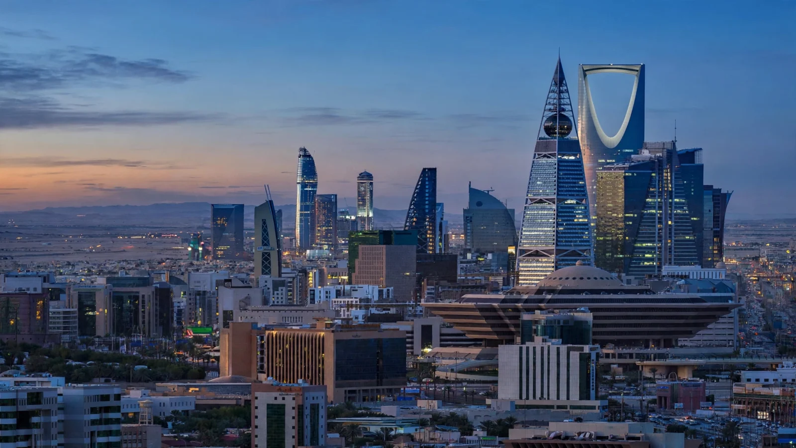 The Riyadh skyline at dusk, with the Kingdom Centre tower and other modern skyscrapers lit.