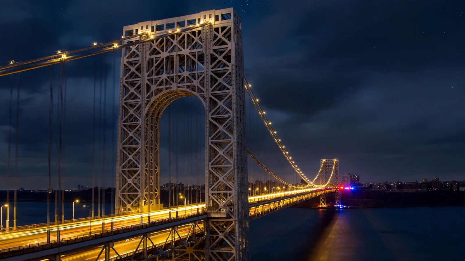Night view of the George Washington Bridge, lit towers and cables, with red and blue emergency lights flashing on the deck in the distance