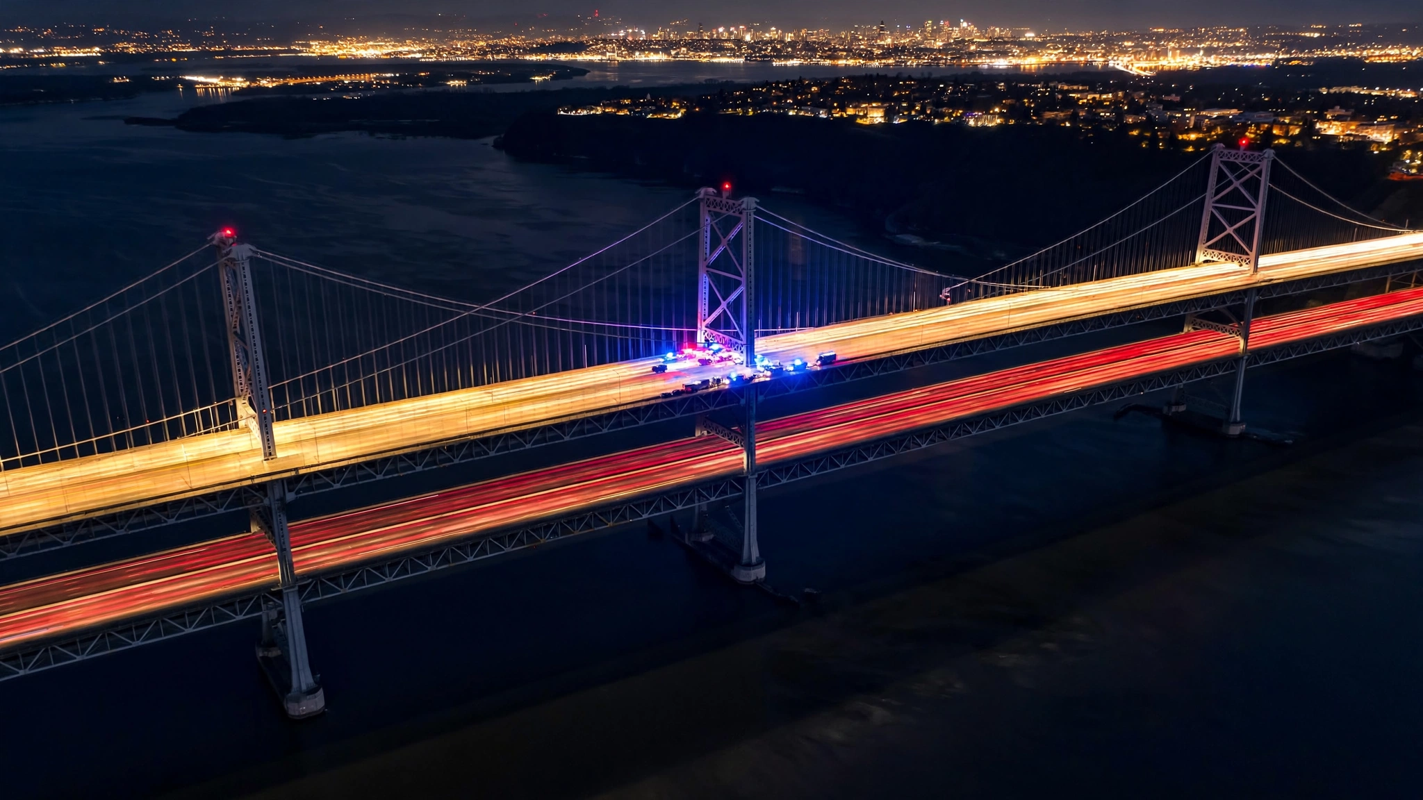 Aerial night view of a suspension bridge over a river, with light trails from traffic and a cluster of blue and red emergency lights mid-span