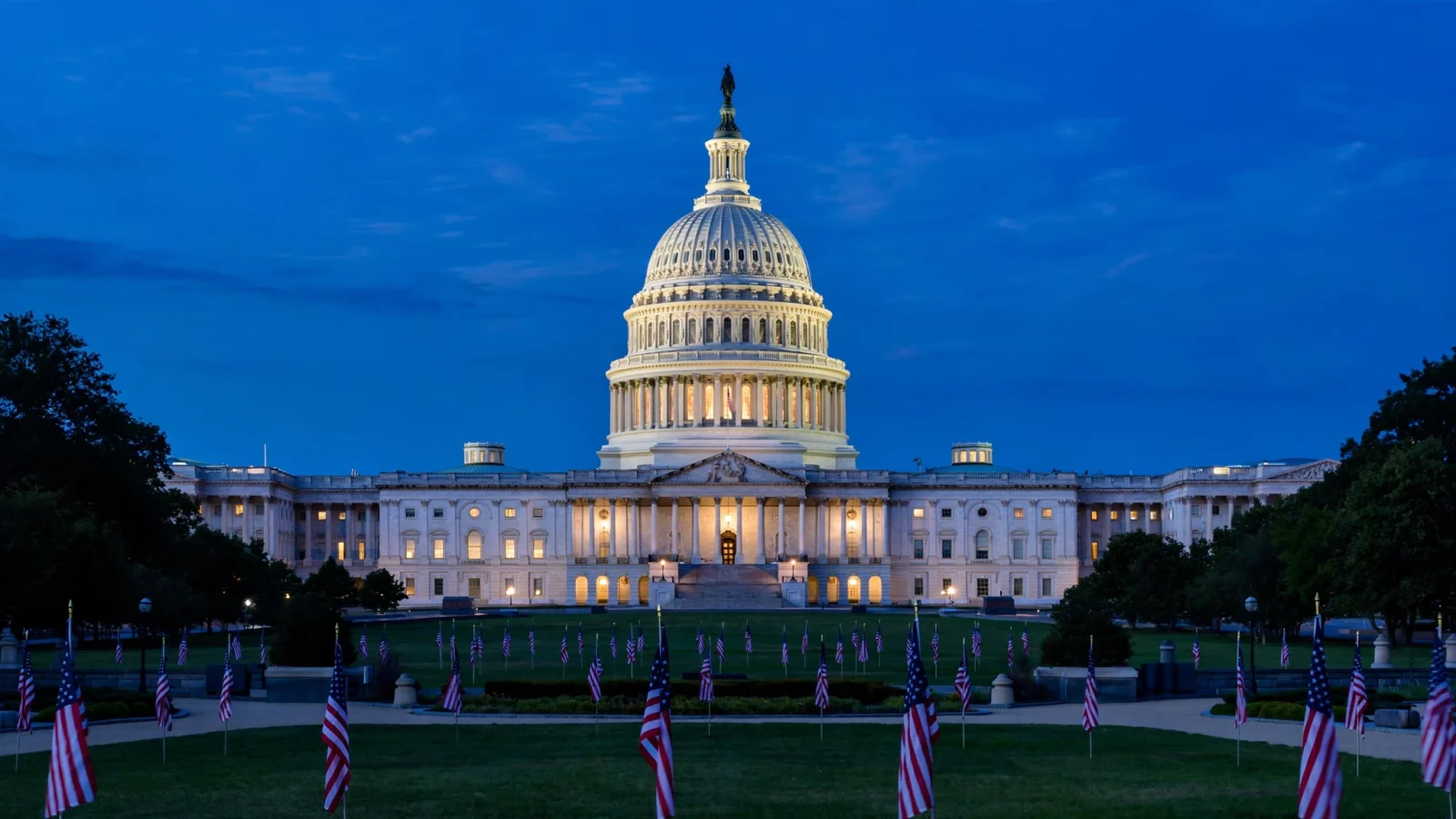 The U.S. Capitol dome lit against a deep blue evening sky, with rows of small American flags on the grounds.